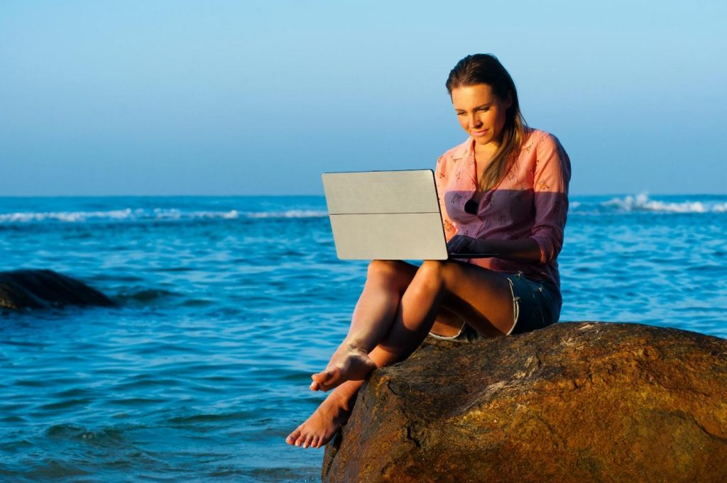 Woman sitting with a laptop on a rock near the sea