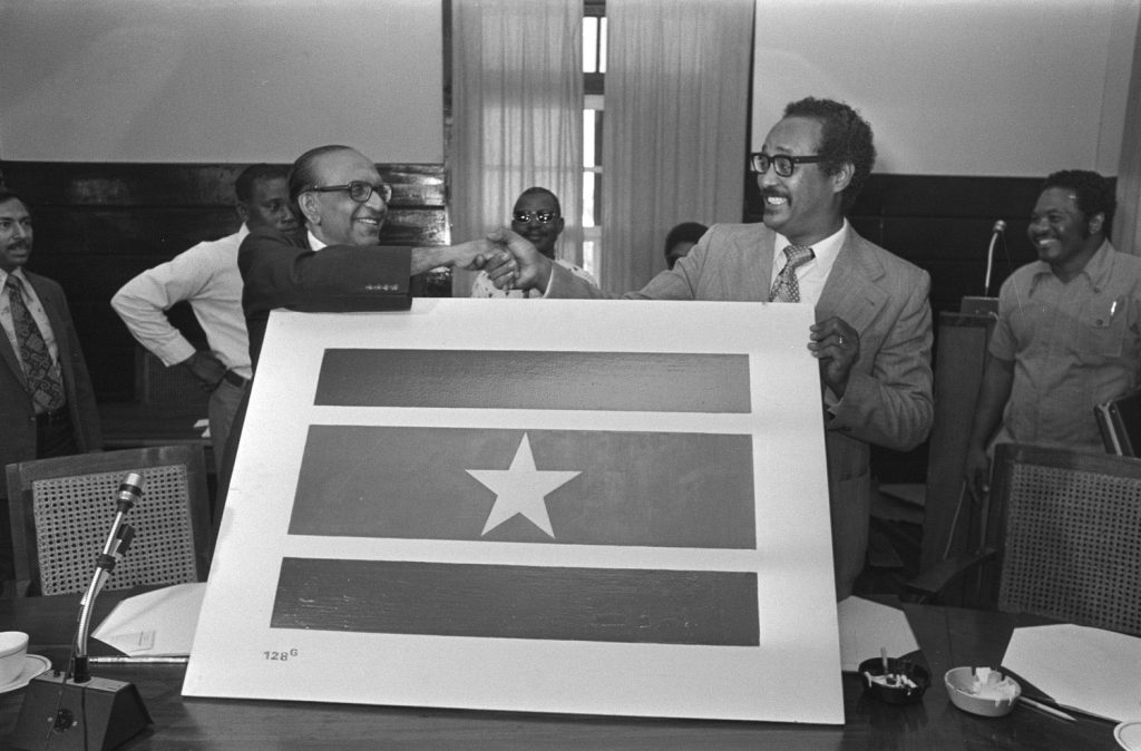 Prime Minister Arron (right) and Opposition Leader Lachmon prepare for independence with the new flag of the Republic of Suriname. Photo courtesy Wikimedia Commons